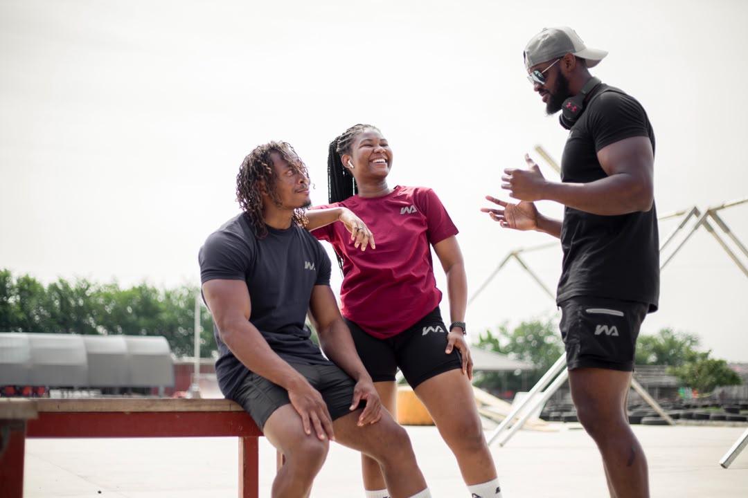 Three people in Warmz tees — charcoal, maroon, and black — laughing together by an outdoor bench.