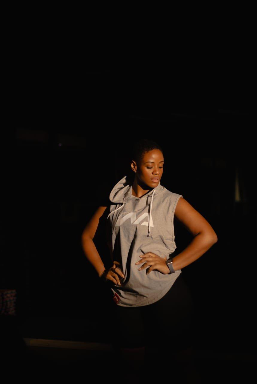 A woman standing with hands on her hips in a grey Warmz sleeveless hoodie, lit in warm amber light in a studio.