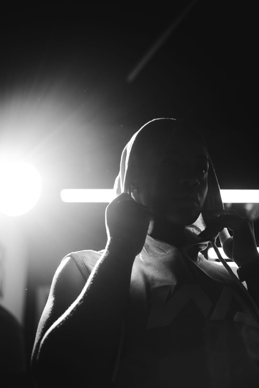 A close-up silhouette of a woman in a grey Warmz hoodie, backlit by a bright studio light, in black and white.