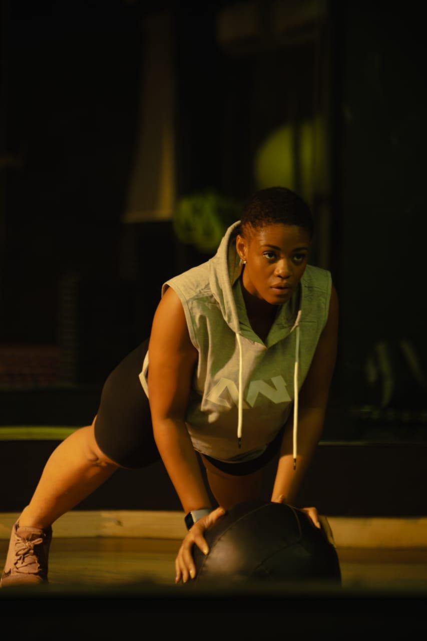 A woman in a grey Warmz sleeveless hoodie and black shorts holding a plank position over a medicine ball, in a dimly lit gym.