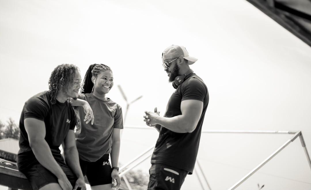 Three people in black Warmz tees laughing together outdoors, a wind turbine visible behind them, in black and white.